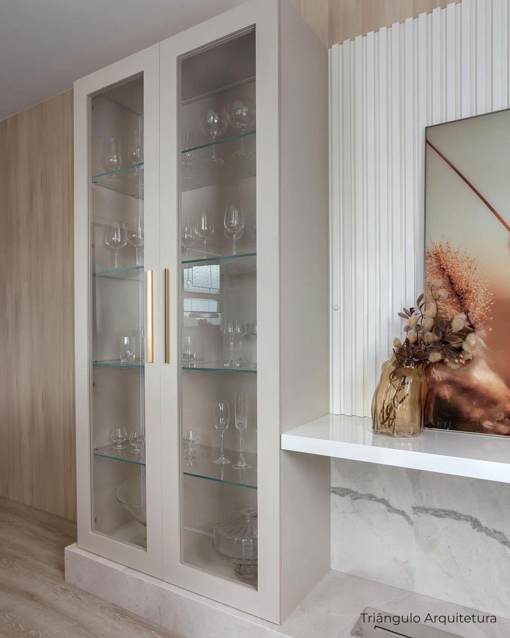 Cristaleira em MDF bege com portas em MDF usinado e vidro transparente, puxador dourado. Prateleiras de vidro e painel ripado em MDF branco ao lado